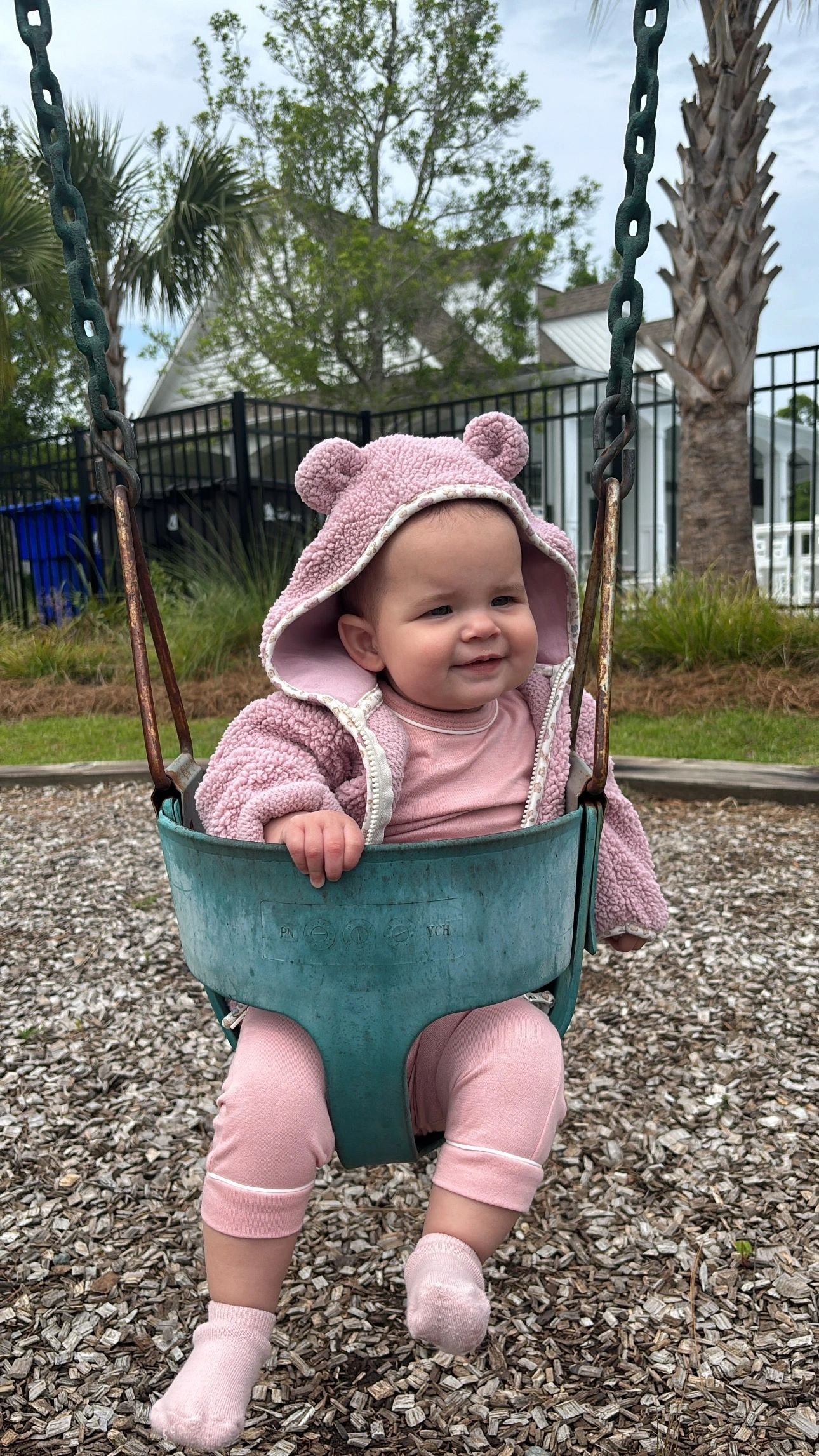 Smiling baby in a pink bear hoodie sitting in a playground swing